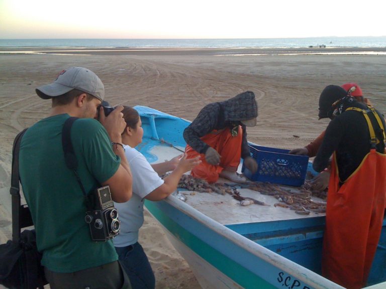 A small group of people gather on and around a fishing boat on an expansive beach. A photographer stands nearby taking their photo.
