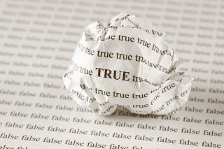 A balled-up piece of paper with the word TRUE typed on it repeatedly sits on a piece of paper with the word FALSE written on it over and over again.