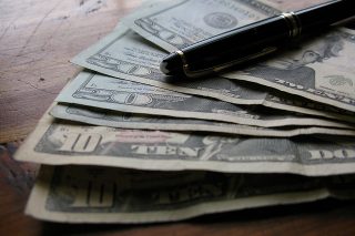 A small pile of $10 and $20 bills sits fanned out on a wooden surface, held down by a black pen.