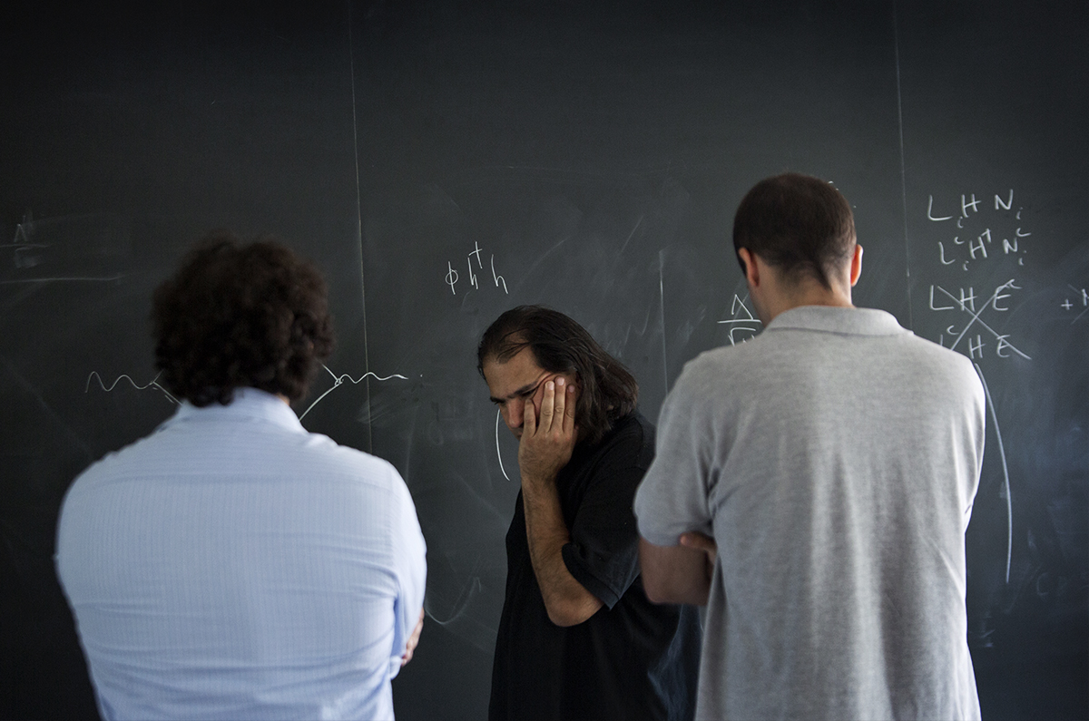 A man standing at a blackboard, caught in thought with his hand to his cheek, as two other men face him.