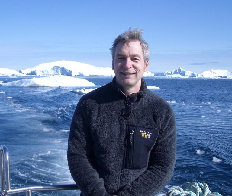 Tim Folger smiles at the camera wearing a navy blue fleece. Behind him, in the wake, dark blue water contains chunks of ice. In the background are large ice structures.