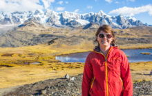 Lizzie Wade in large sunglasses and a red walking jacket with yellow trim smiles at the camera. In the background is a range of snow-topped mountains with flatter grasslands and a small lake just behind her.