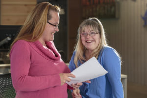 Two women stand close to each other. One in a pink sweater and glasses is holding papers and smiling at the other woman. She is wearing glasses and a blue top. She is smiling in the direction of the camera and has facial features commonly seen in Down syndrome.