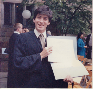 A man wearing a graduation gown smiles while holding up a diploma.