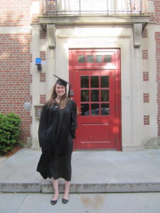 A woman wearing a graduation cap and gown stands in front of a brick building with a red door.