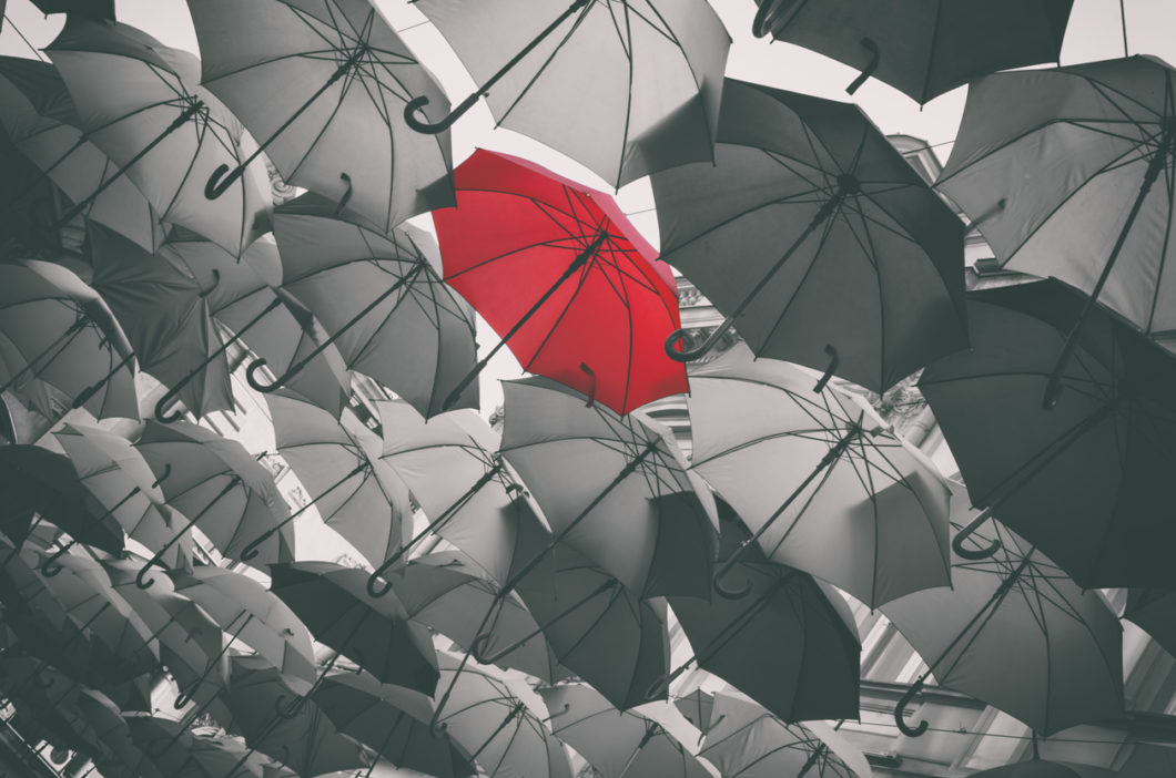 A collection of umbrellas in greyscale, with one red umbrella in the center of the grouping.