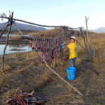 Photo of a smiling kid, who is hanging some fish on a wooden structure. There is water nearby and mountains on the background.