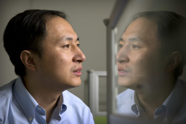 Photo of Chinese scientist He Jiankui looking away from the camera; we can see his reflection on a piece of glass.