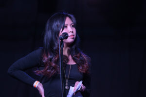 Eli Chen standing in front of a microphone, in a dark background, she is illuminated by the room lights.