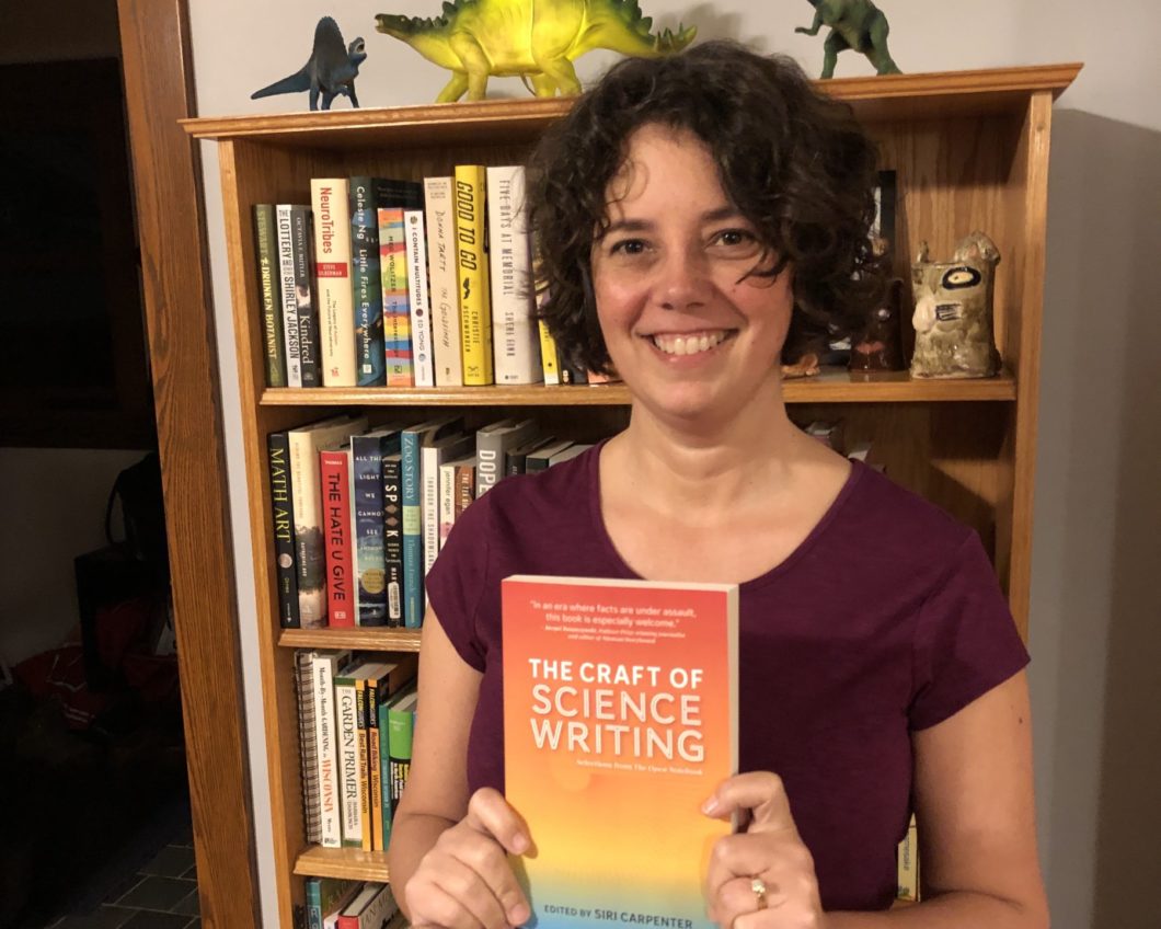 Siri Carpenter holding a copy of The Craft of Science Writing, standing in front of a bookcase.