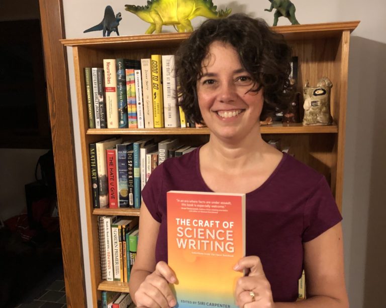 Siri Carpenter holds a copy of The Craft of Science Writing, standing in front of a bookcase.