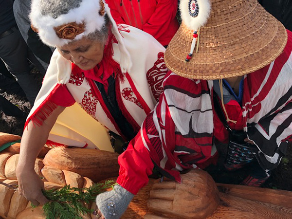 Two people in red and white attires, leaning forward with their arms extended to reach a wooden artifact.
