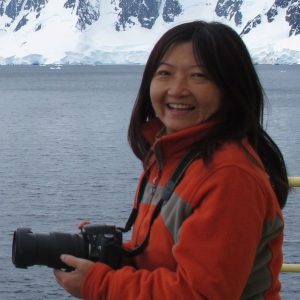 A woman looks directly into the camera, while holding a camera on her hands, at the height of her chest. Behind her, glaciers rise in front of a lake or the ocean.