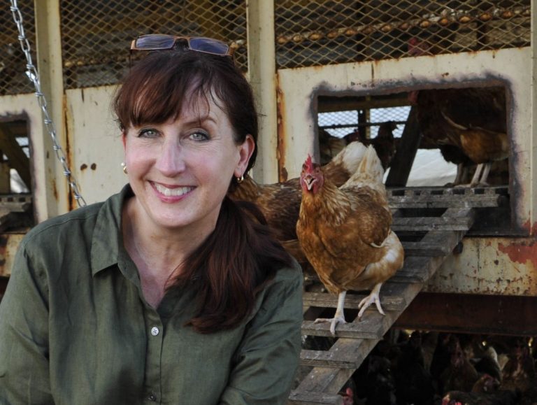 A woman looks into the camera, smiling, sitting in front of a chicken coop and a number of hens.