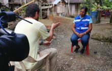 Juan Miguel Álvarez interviewing a woman. They sit across each other on an unpaved street surrounded by wooden homes in Colombia. Behind Álvarez, there's a camera lens pointing in the direction of the woman he interviews.