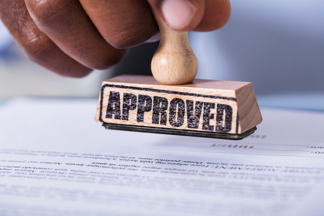 A hand is holding a wooden stamp with the word "APPROVED" printed on it over an unreadable document.