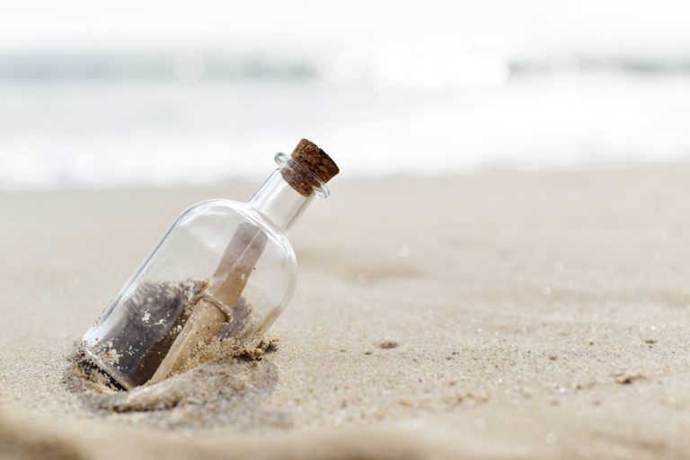 A glass bottle with a rolled-up message inside, wedged in the sand on a beach.
