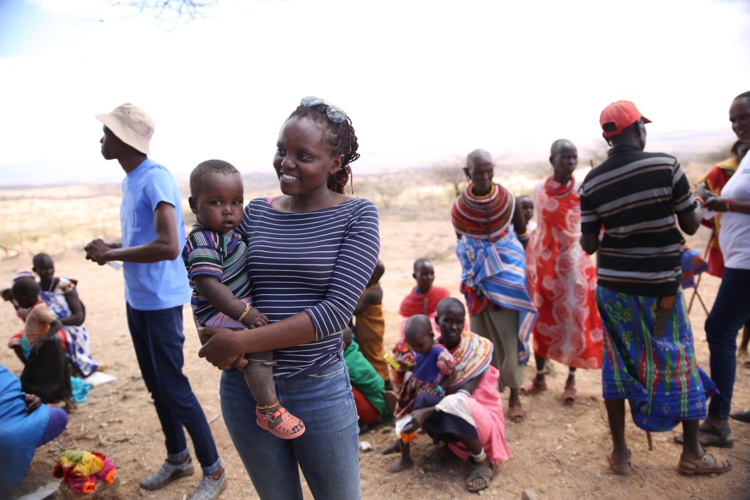Verah stands outdoors holding a small child while speaking to someone out of the frame. Behind her is a large group of people dressed in colorful clothing.