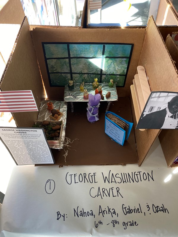 Peeps diorama showing inventor George Washington Carver working in his lab, with several types of vegetables next to him, and a printed copy of his most popular bulletin, How to Grow the Peanut and 105 Ways of Preparing it for Human Consumption. 