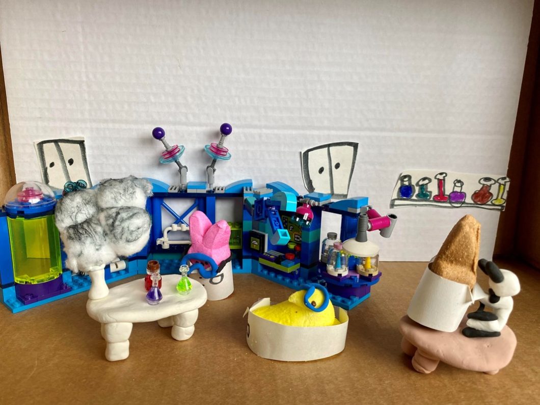 Peeps diorama showing an explosion inside a lab, while a pair of distracted scientists, wearing googles and lab coats, look into a microscope without realizing what's going on. 