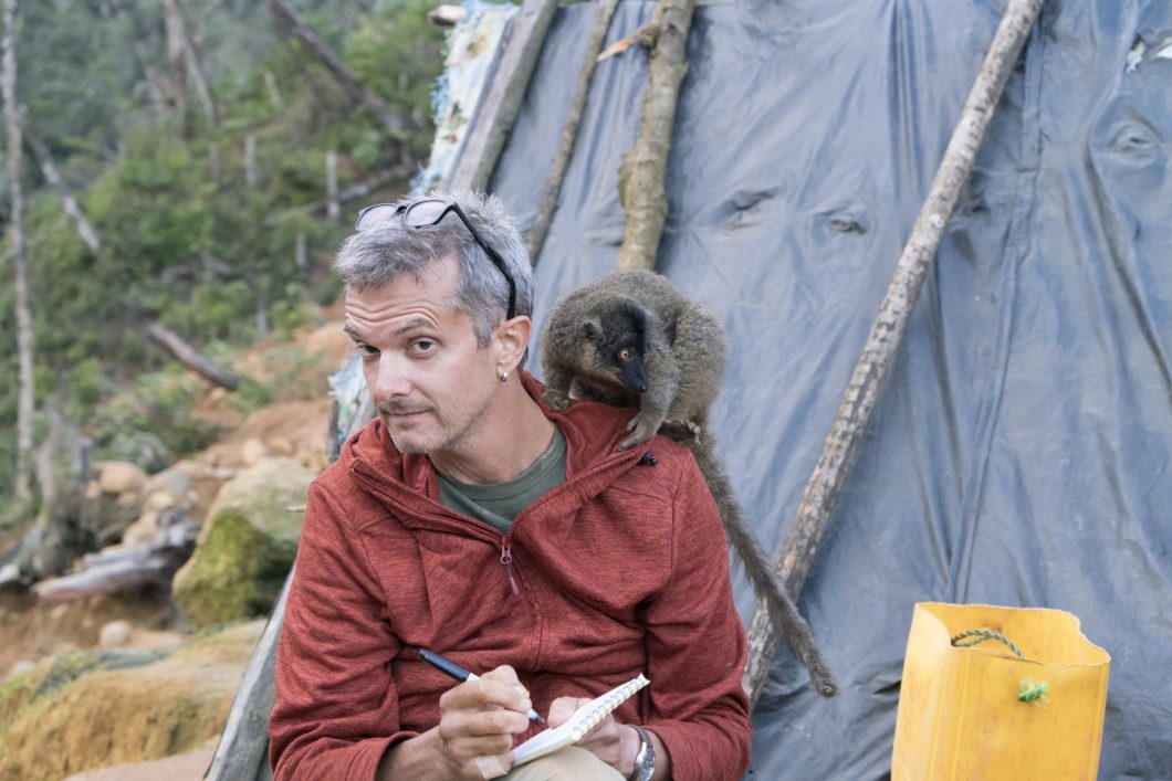 Paul Tullis sitting in front of a tent with a pen and notebook. There is a lemur on his left shoulder.