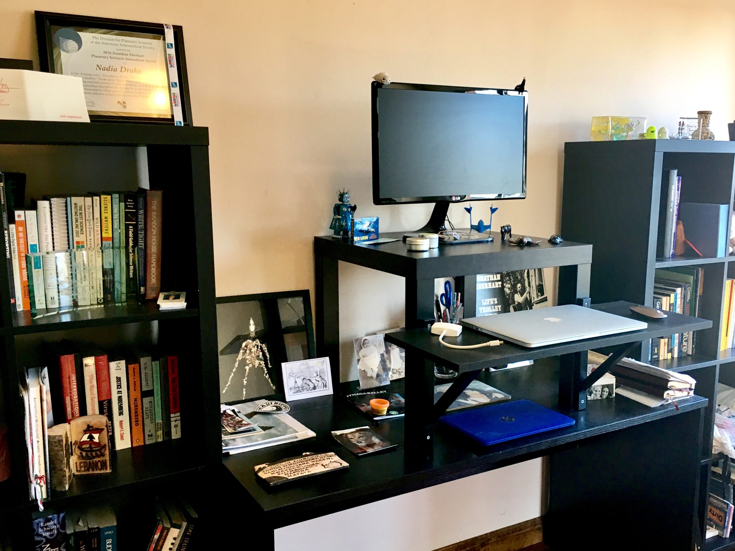A black standing desk with laptop, monitor, and miscellaneous objects, and bookshelves on both sides.
