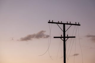 A silhouette of a telegraph pole with broken wires against a dimly lit sky with small clouds.