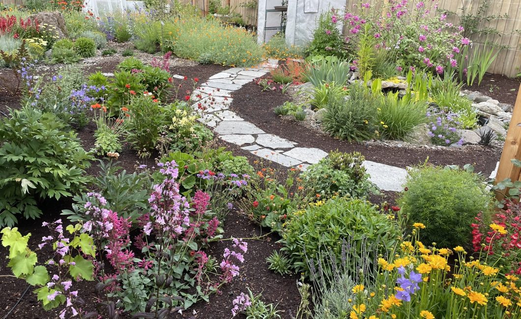 A lush, beautifully tended garden with flowers of many different colors. A path curves through the middle of the image toward a small greenhouse.