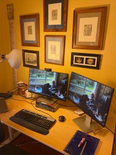 Desk with two computer monitors, a keyboard, and a variety of odds and ends. The desk is up against a yellow wall that has several framed images hanging on it.
