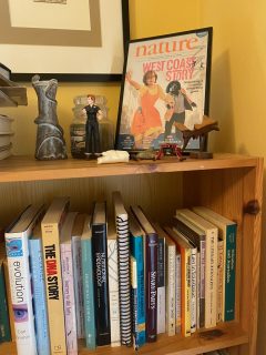 A closeup shot of a bookshelf containing books on science and journalism. On the top shelf there are trinkets and a framed faux Nature magazine cover with Rosie holding a dog's hand. The headline is "West Coast Story."