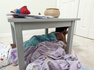 Ein Kind kuschelt sich mit einer lila Decke unter einen Tisch und liest ein Buch.