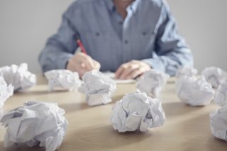A bunch of crumpled paper balls sit on a desk in the foreground. In the background, a writer sits at the desk with pencil and paper, trying again.