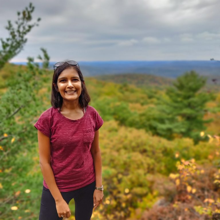A woman stands on a rise facing the camera. In the background there is a long view of autumnal trees in a mountain valley.