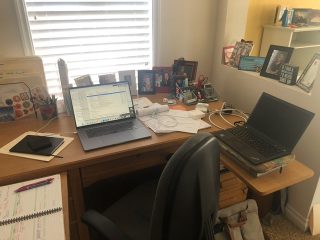 A desk, which holds two computers, a planner, notepads, and framed portraits, sits in front of a sunny window.