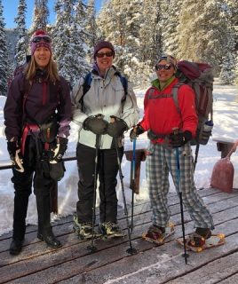 Three friends wearing backpacks and snowshoes stand on a rooftop surrounded by a winter wonderland.