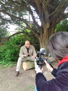 Khadka sits in front of a camera under a large tree in London's Yeading Walk while reporting on the failures of several reforestation programs across the globe.