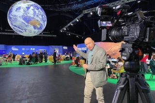 BBC global environment correspondent Navin Singh Khadka stands in front of a large video camera on a tripod while reporting on the United Nations Climate Change Conference in 2021. In the background attendees sit at tables and mingle. A large globe, or photo of Earth, hangs from the ceiling.