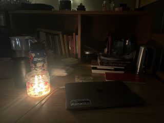 A laptop computer, books, and a burning candle, among various other items, sit on a desk in a dark room.